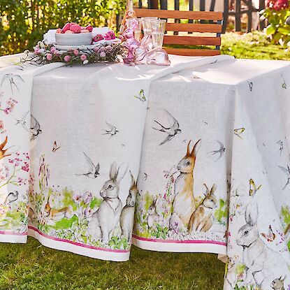 Oster-Tischdecke mit Hasen- und Vogelmotiven, fr&uuml;hlingshaft gedeckter Gartentisch mit Deko und Geb&auml;ck.