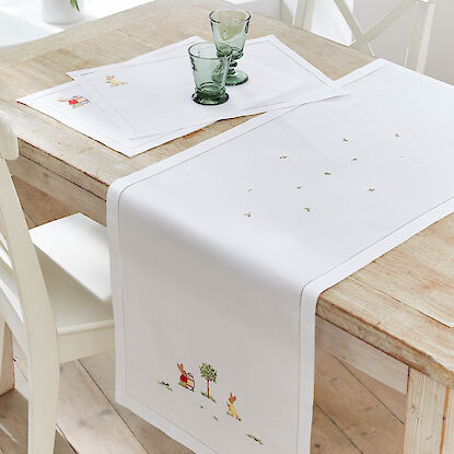 Wei&szlig;e Tischw&auml;sche mit feiner Hasenstickerei, bestehend aus Tischl&auml;ufer und Platzsets auf hellem Holztisch.