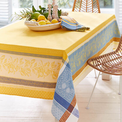 Gelbe Tischdecke mit blauem Bord&uuml;renmuster, gedeckter Tisch mit Zitronen, Kerzen und Servietten im hellen Essbereich.