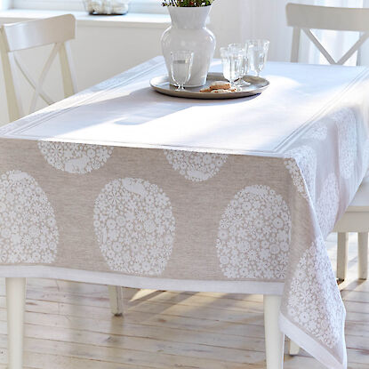 Helle Tischdecke mit dezentem Hasenmotiv, gedeckter Tisch mit Vase, Gl&auml;sern und Tablett in lichtdurchflutetem Raum.
