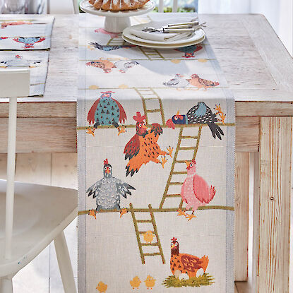 Tischl&auml;ufer mit bunten H&uuml;hner- und K&uuml;kenmotiven, fr&uuml;hlingshaft gedeckter Holztisch mit Kuchen und Geschirr.