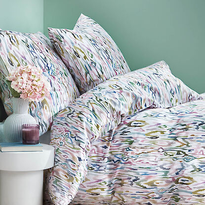 Gemusterte Bettw&auml;sche in Pastellfarben, modern arrangiertes Bett mit Kissen, Vase und Nachttisch vor gr&uuml;ner Wand.