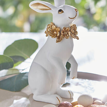 Wei&szlig;e Hasenfigur mit goldenem Blumenkranz, dekorativ arrangiert auf Tablett mit Ostereiern im Fr&uuml;hlingslicht.