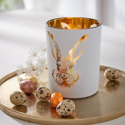 Kerzenglas mit goldenem Hasenmotiv, brennende Kerze auf Tablett mit dekorativen Ostereiern in warmem Licht.