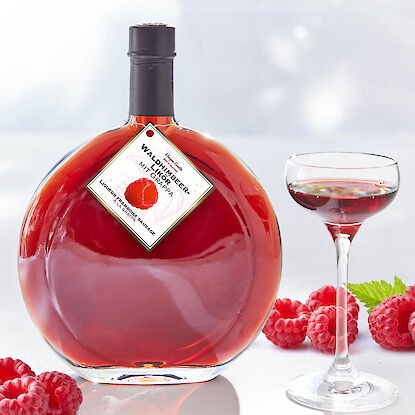 Himbeerlik&ouml;r in runder Glasflasche mit gef&uuml;lltem Lik&ouml;rglas und frischen Himbeeren auf hellem Hintergrund.