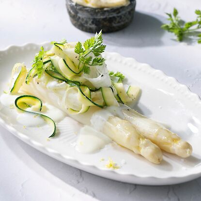Wei&szlig;er Spargel mit Zucchinistreifen und heller Sauce auf Teller, garniert mit Kr&auml;utern