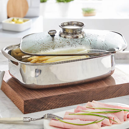 Edelstahl-Spargelbr&auml;ter mit Glasdeckel auf Holzbrett, wei&szlig;er Spargel im Topf, Schinken auf Teller