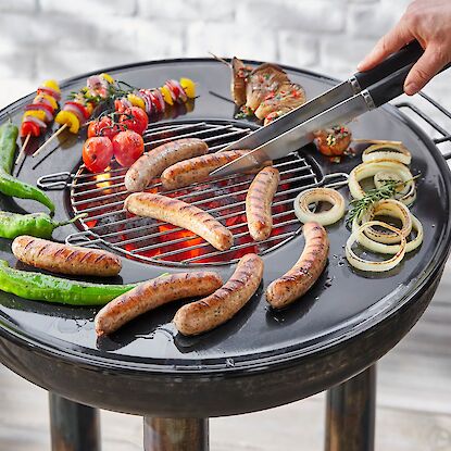 Holzkohlegrill mit Bratw&uuml;rsten, Paprika, Zwiebeln und Gem&uuml;sespie&szlig;en, Person wendet W&uuml;rste mit Grillzange