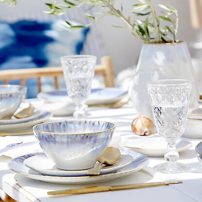 Sommerlich gedeckter Tisch mit Geschirr, Glas und Vase mit Olivenzweig im Garten
