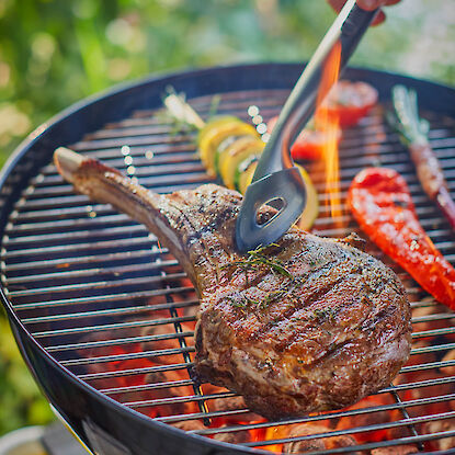 Tomahawk-Steak auf Holzkohlegrill mit Grillzange, Gem&uuml;se und Flammen