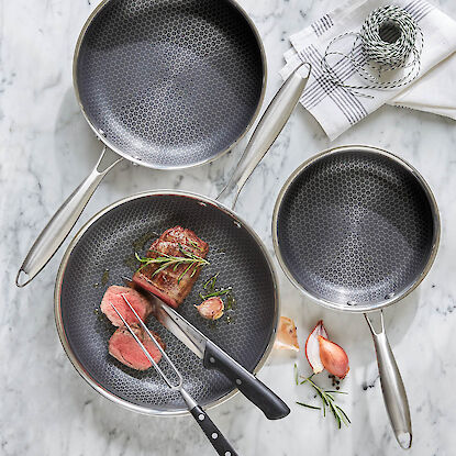 Edelstahlpfannen mit Wabenstruktur-Beschichtung, Steak und K&uuml;chenutensilien auf Marmortisch