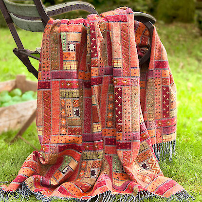 Gemusterte Decke in warmen Rot- und Oranget&ouml;nen &uuml;ber Gartenstuhl im Gr&uuml;nen drapiert