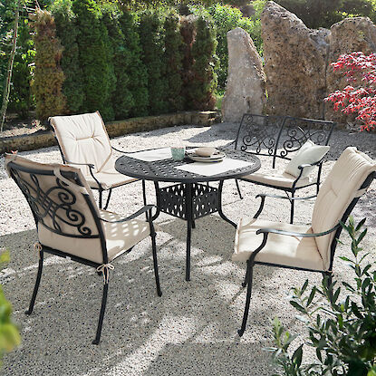 Gartentisch aus Metall mit gepolsterten St&uuml;hlen auf Kiesfl&auml;che, umgeben von Pflanzen und Naturstein