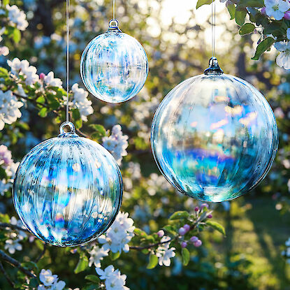 H&auml;ngende Glaskugeln in Blau schimmernd zwischen bl&uuml;henden Zweigen im sonnigen Garten