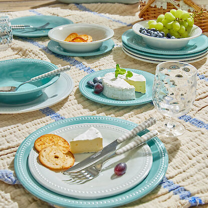 Picknickgedeck mit t&uuml;rkisfarbenem Geschirr, K&auml;se, Obst und Gl&auml;sern auf Decke im Freien