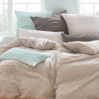 Bettw&auml;sche aus Leinen in nat&uuml;rlichen Beige- und Graut&ouml;nen, kombiniert mit Kissen in sanften Pastellfarben, locker auf einem Bett arrangiert