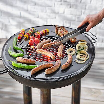 Holzkohlegrill mit Bratw&uuml;rsten, Paprika, Zwiebeln und Gem&uuml;sespie&szlig;en, Person wendet W&uuml;rste mit Grillzange