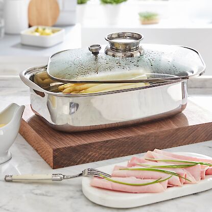 Edelstahl-Br&auml;ter mit Glasdeckel und Spargel, serviert auf Holzbrett, daneben Sauce und Schinken auf Teller