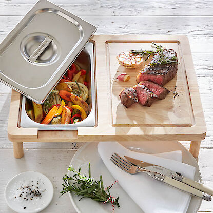 Schneidebrett mit integriertem Edelstahlbeh&auml;lter, serviert mit Steak, Ofengem&uuml;se und Knoblauch, daneben Teller und Besteck