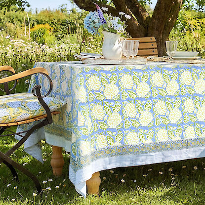 Gartentisch mit gemusterter Tischdecke, gedeckt mit Geschirr und Blumen, unter Baum im sommerlichen Garten