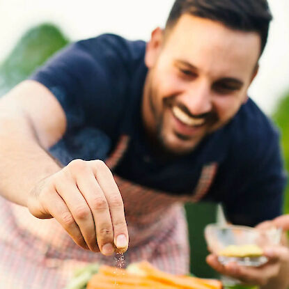 Mann w&uuml;rzt Fleisch auf Grill mit Gew&uuml;rzen, l&auml;chelnd beim Grillen im Garten mit Gem&uuml;se im Hintergrund