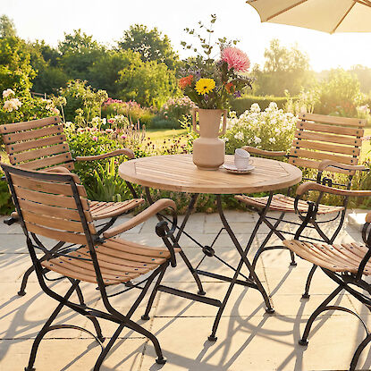 Gartenm&ouml;bel-Set aus Holz und Metall mit rundem Tisch und St&uuml;hlen, dekoriert mit Blumen auf sonniger Terrasse