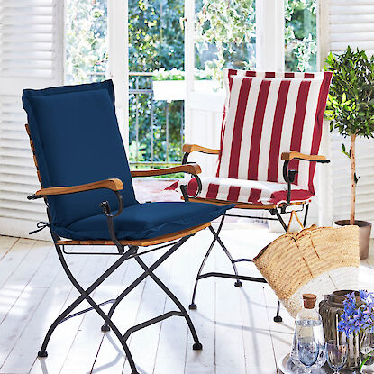 Klappbare Gartenst&uuml;hle mit Polstern in Blau und Rot-Wei&szlig; gestreift auf heller Terrasse, dekoriert mit Tablett und Korb