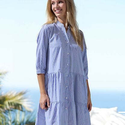 Damenkleid in Blau-Wei&szlig; gestreift mit Knopfleiste und lockerem Schnitt, getragen auf sonniger Terrasse mit Meerblick