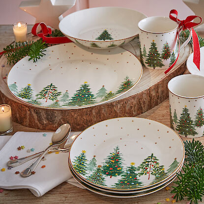 Vaisselle de Noël avec motif de sapin, entourée de bougies et de branches de sapin.