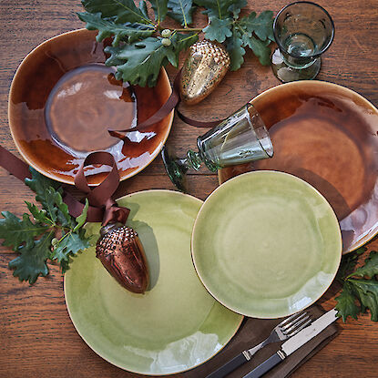 Table automnale dressée avec des assiettes dans des tons bruns et verts, glands et verre.