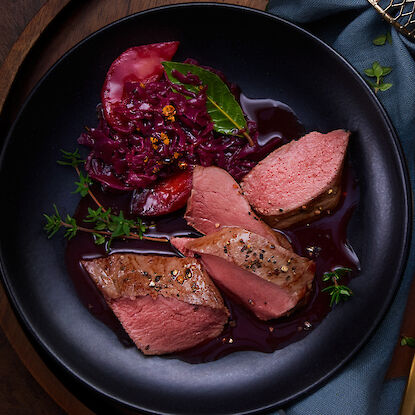 Perfekt gebratenes Fleisch mit Rotkohl, Apfelscheiben und Kräutern auf einem schwarzen Teller.