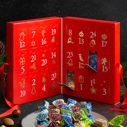 Roter Adventskalender mit geöffneten Türchen, daneben eine Platte mit verpackten Pralinen.