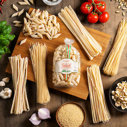 verschiedene Pasta-Sorten auf einem Holzbrett, umgeben von frischen Zutaten wie Tomaten, Knoblauch und Basilikum.