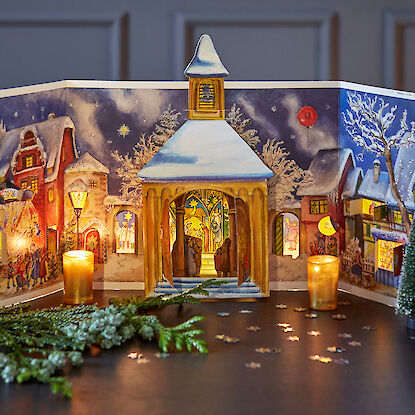 Aufgestellter Adventskalender in einer winterlichen Dorfkulisse, umgeben von Kerzen und Miniaturtannen