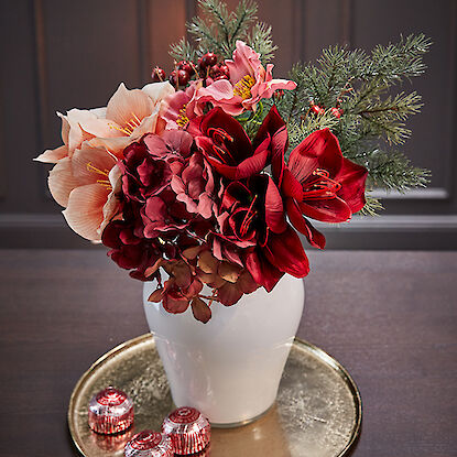 Festlicher Blumenstrauß in Rot- und Rosatönen, arrangiert in einer weißen Vase, auf einem goldenen Tablett mit dekorativen Pralinen.