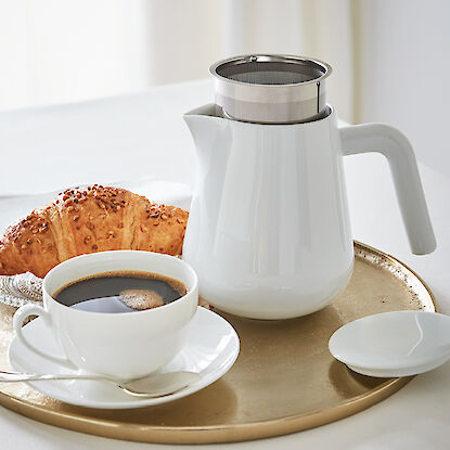 Frühstück mit weißer Teekanne, Tasse Kaffee und Croissant auf goldenem Tablett