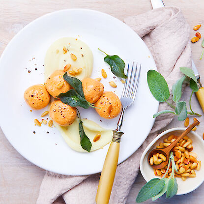 Elegantes Gericht mit Lachsklösschen, Salbeiblättern, Pinienkernen und Püree auf einem weißen Teller, dekoriert auf Holztisch.