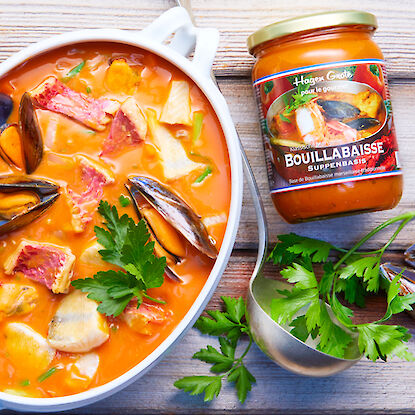 Bouillabaisse mit Fisch und Meeresfrüchten im Topf, daneben Suppenbasis im Glas und frische Petersilie.