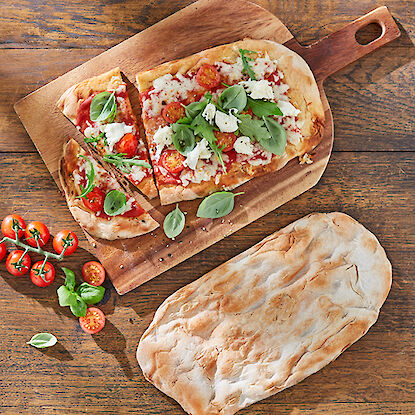 Pinsa fraîche avec tomates, basilic et fromage sur une planche en bois, accompagnée d’une focaccia entière.