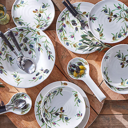 Service en céramique au motif d’olivier, avec assiettes, bols et couverts sur table en bois.