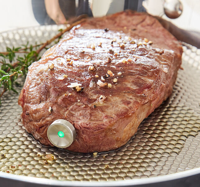 Steak mit digitalem Fleischthermometer auf einer gerillten Pfanne, verfeinert mit Gewürzen und frischen Kräutern.