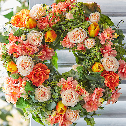 Ein bunter Blumenkranz mit Rosen, Tulpen und grünen Blättern hängt vor einem hellen, hölzernen Hintergrund