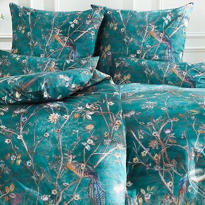 Elegantes Schlafzimmer mit einem Bett mit dunkelgrün-blauer Bettwäsche mit einem auffälligen Dessin mit Pfauen, Zweigen mit unterschiedlichsten Blüten daneben graziele Nachttische in Gold und Weiß mit Lampen und Vasen mit Blumenstrauß