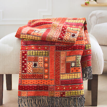 Rot-gelbes Plaid im Patchwork-Stil auf einem mit weißem Stoff bezogenem rechteckigem Hocker in einem hellen Raum mit Kassettenwand im Hintergrund