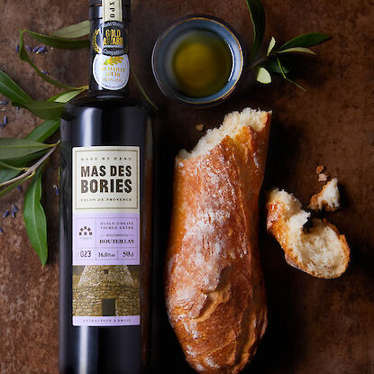 Edles Olivenöl Mas des Bories aus der Provence, serviert mit knusprigem Baguette und Olivenzweigen.