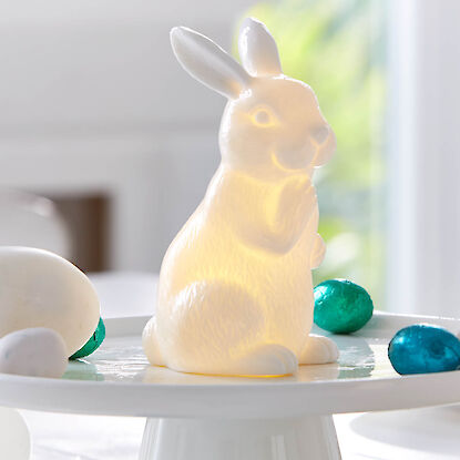 Lapin de Pâques lumineux en porcelaine sur un présentoir blanc, entouré d'œufs décoratifs.