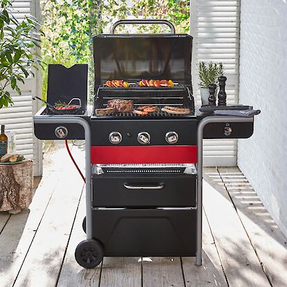 Gasgrill mit drei Brennern, Seitenkochplatte und Grillgut auf einer Terrasse.