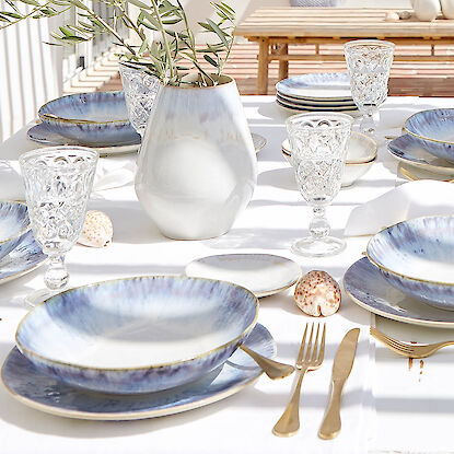 Sommerlich gedeckter Tisch mit blau-weißem Geschirr, Goldbesteck und Vase mit Olivenzweig