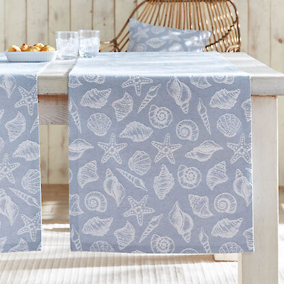 Tischläufer in Hellblau mit Muschelmotiv auf Holztisch, dazu passende Deko und Korbstuhl