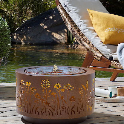 Runder Gartenbrunnen mit floralem Muster auf Holzdeck neben Liegestuhl und Naturteich.